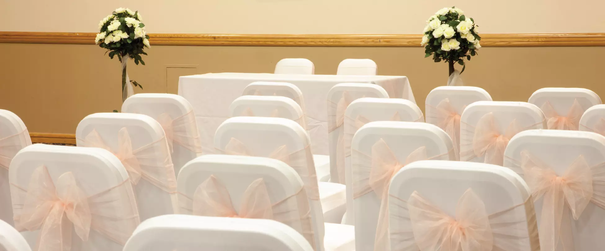Chairs set up for a wedding ceremony with white covers and peach ribbons. They are facing a table set up for the ceremony covered in white with a bouquet of white flowers to either side..
