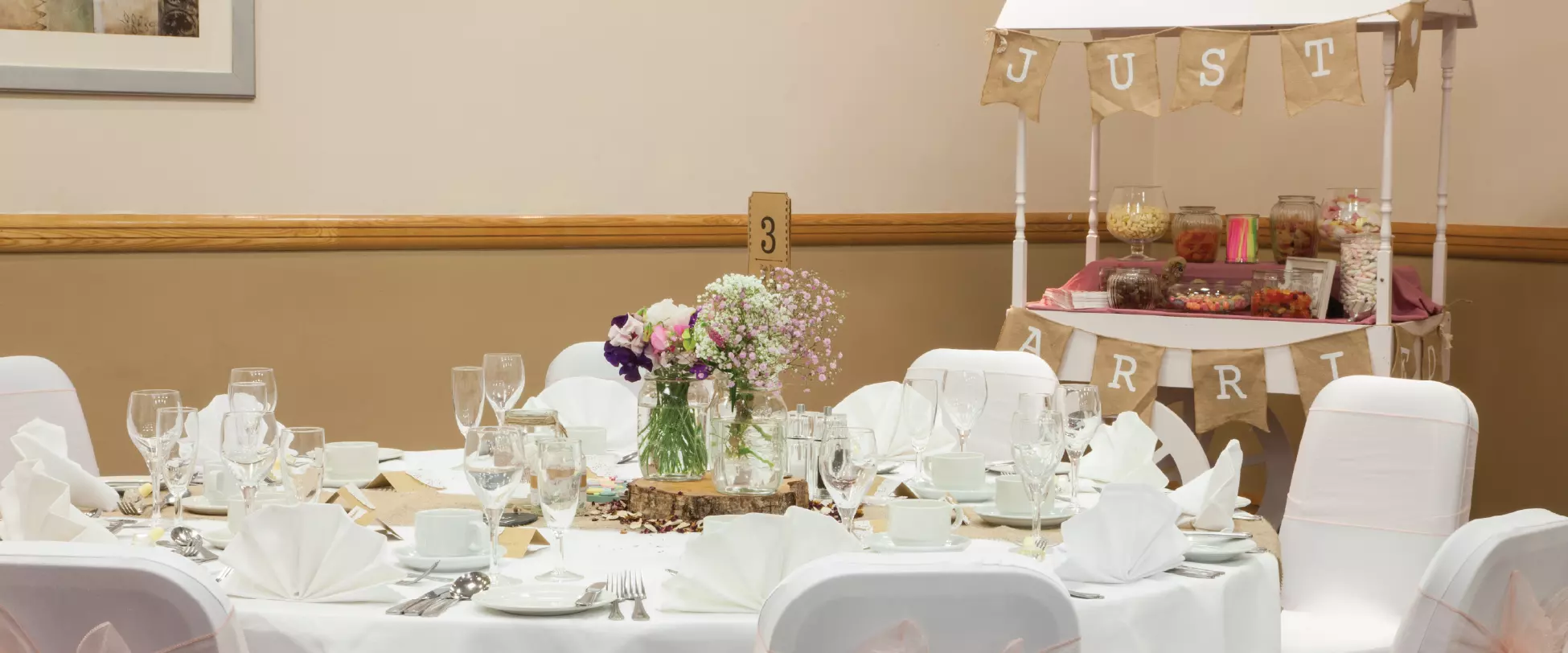 A table set up for a wedding breakfast with white table and chair covers. There's a stall in the background with sweets.